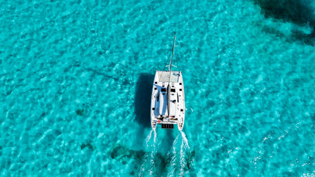 Aerial view of a white catamaran cruising over crystal-clear turquoise tropical water with twin wakes trailing behind