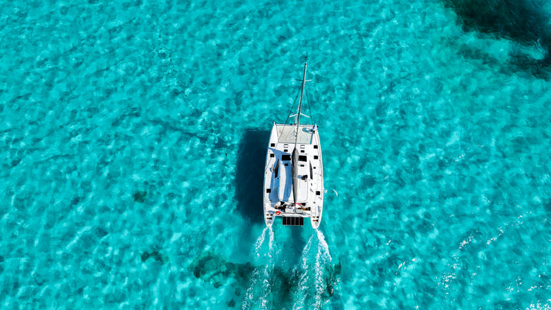 Aerial view of a white catamaran cruising over crystal-clear turquoise tropical water with twin wakes trailing behind