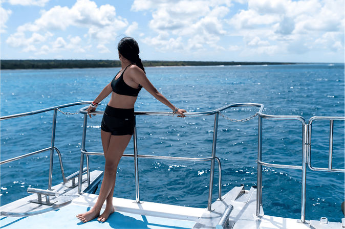 Woman in black swimwear standing barefoot on a boat deck, leaning on stainless-steel railing and gazing at a sun-soaked turquoise ocean and distant tropical coastline under a blue sky with fluffy clouds.