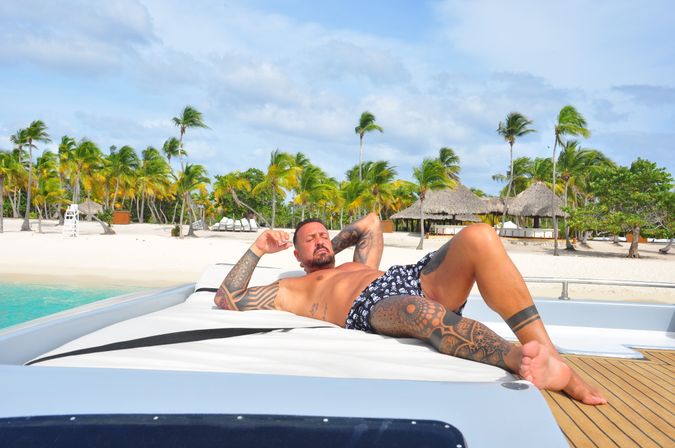 Tattooed man in swim trunks lounging on a yacht sunbed off a tropical white-sand beach with palm trees, turquoise water and thatched beach huts