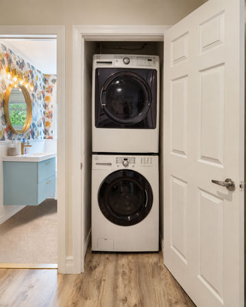Stacked front-loading washer and dryer in a narrow home laundry closet with light wood flooring, open white door leading to a colorful floral wallpaper bathroom with a blue floating vanity and round gold mirror.