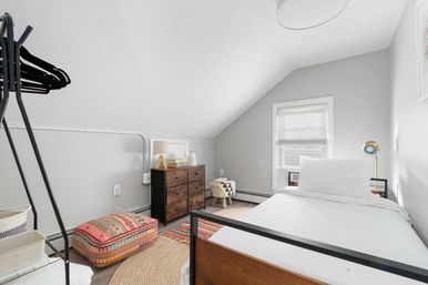 Cozy attic bedroom with sloped ceiling, single white bed with wooden frame, dark wood dresser, patterned boho floor pouf and woven rugs under a sunlit window
