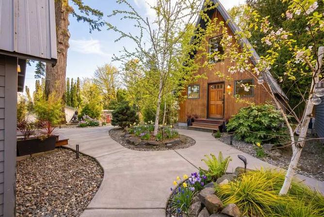 Cozy A-frame wooden cabin with a curved concrete walkway through a landscaped front yard featuring spring blossoms, rock beds, and colorful tulips and irises.