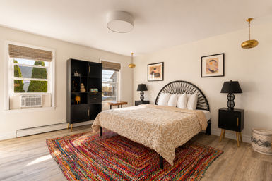 Sunlit cozy modern bedroom interior with a black rattan headboard, beige textured bedspread, colorful woven area rug, matching black nightstands and lamps, tall black shelving unit, bamboo window shades, brass pendant lights and light wood floors.