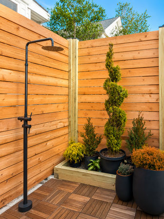 Sleek black outdoor rain shower beside a wooden privacy fence corner with potted topiary and evergreen plants on a tiled backyard patio