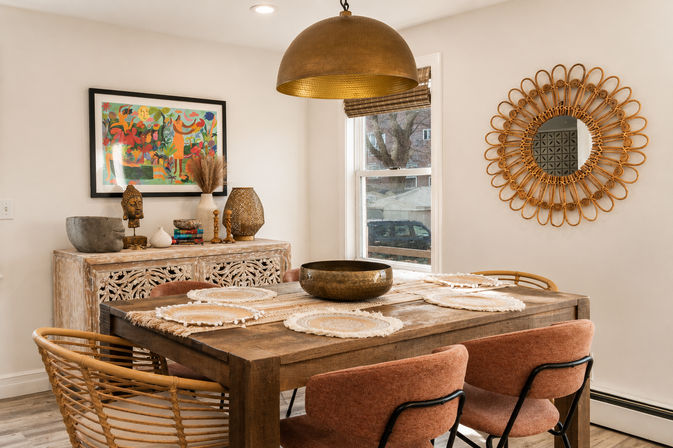 Sunlit boho dining room with a rustic wooden table topped with woven placemats and a hammered brass bowl, rattan and upholstered chairs, large brass pendant light, carved whitewashed sideboard with colorful framed art and decorative sculptures, and a round rattan mirror on a neutral wall.