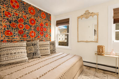 Sunlit bohemian bedroom with a vibrant orange-and-navy floral tapestry above a beige pompom quilt, patterned black-and-white throw pillows, antique-style mirror, wicker basket and pink glass vase on a small side table.