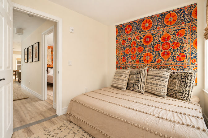 Cozy boho guest bedroom with vibrant orange-and-blue floral tapestry above a pom-pom trimmed neutral daybed, patterned throw pillows, hardwood floors, and an open doorway to a hallway.