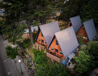 Aerial view of cozy wooden A-frame cabins with gray metal roofs nestled among tall evergreens and a landscaped rock garden beside a quiet streetlamp