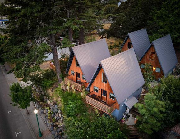 Aerial view of cozy wooden A-frame cabins with gray metal roofs nestled among tall evergreens and a landscaped rock garden beside a quiet streetlamp