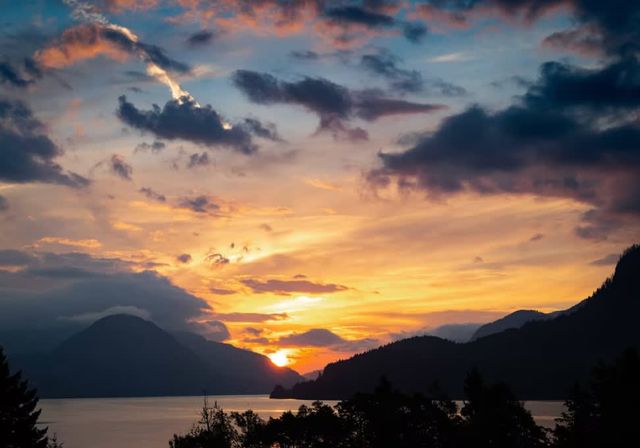 Vibrant mountain-lake sunset with the sun dipping behind silhouetted peaks, golden-pink clouds reflected on calm water