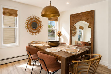 Sunlit boho-chic dining room with a rustic wooden table, woven placemats, rattan and upholstered chairs, large carved wood mirror, round rattan wall mirror and brass pendant light