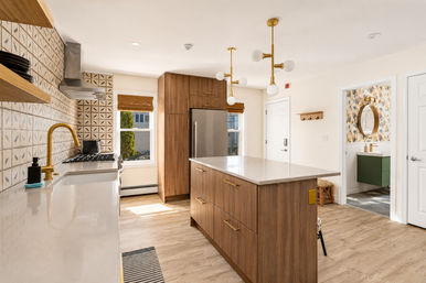 Bright modern residential kitchen with a walnut island and quartz countertop, brass faucet and hardware, patterned tile backsplash, stainless refrigerator, mid-century brass pendant lights, light wood floors, and a colorful powder room visible