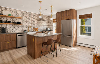 Sunlit modern suburban kitchen with walnut cabinets, patterned tile backsplash, stainless steel fridge and dishwasher, quartz island with brown leather bar stools, brass faucet and globe pendant lights.