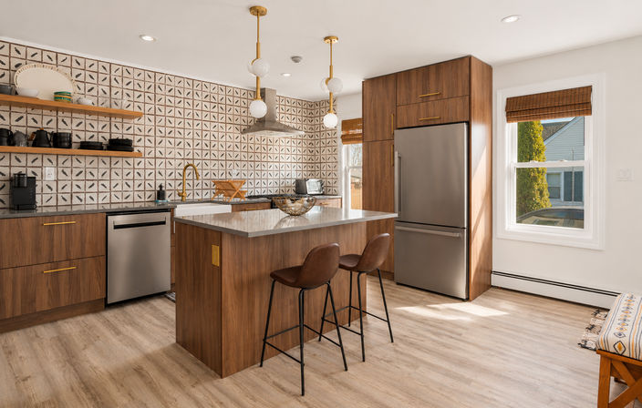 Sunlit modern suburban kitchen with walnut cabinets, patterned tile backsplash, stainless steel fridge and dishwasher, quartz island with brown leather bar stools, brass faucet and globe pendant lights.