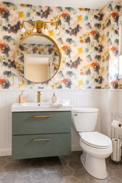 Bright powder room with colorful watercolor floral wallpaper, round brass mirror and light fixture, sage-green floating vanity with brass faucet and pulls, white sink and toilet, and gray hexagon tile floor.