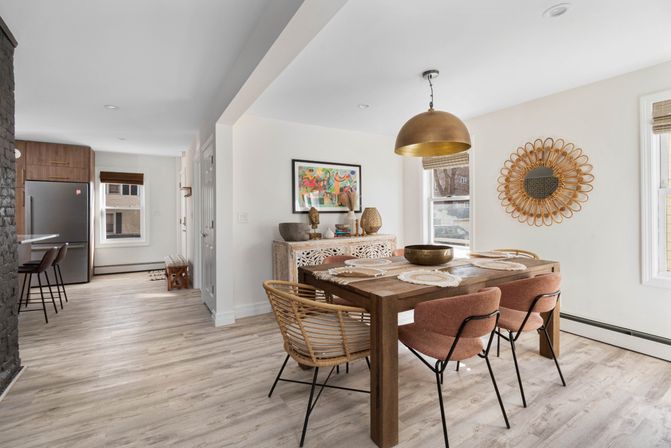 Bright open-concept dining area with a wooden farmhouse table, rattan and blush upholstered chairs, brass dome pendant light, sunburst wall mirror and carved wood sideboard, light wood flooring and stainless refrigerator visible in the adjoining kitchen.