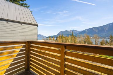 Sunlit wooden balcony railing with panoramic mountain and lake view, forested shoreline and clear blue sky streaked with contrails