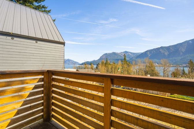 Sunlit wooden balcony railing with panoramic mountain and lake view, forested shoreline and clear blue sky streaked with contrails