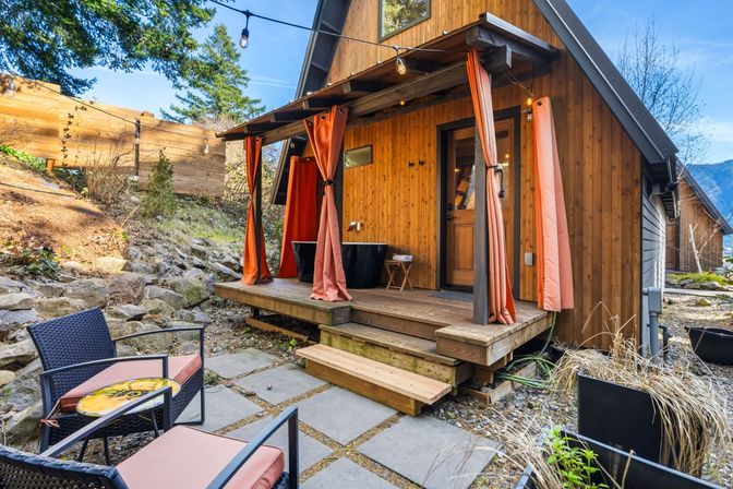 Cozy wooden mountain cabin porch with orange curtains and string lights, a black soaking tub on the deck, cushioned patio chairs and a small table on stone pavers amid rocky landscaping.