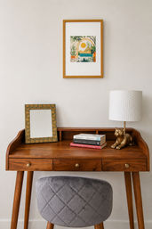 Cozy home office nook with a mid-century wooden writing desk, framed tropical print on the wall, tabletop mirror, stacked books and a whimsical gold cat table lamp with a white textured shade, paired with a gray quilted stool.