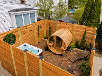 Cozy suburban backyard patio in a private fenced yard featuring a wooden barrel sauna, bubbling hot tub, lounge chairs, potted plants and string lights.