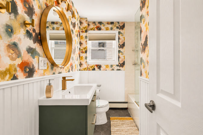 Vibrant cottage-style bathroom with watercolor floral wallpaper, round gold mirror, green vanity with gold faucet, white beadboard and tub-shower.