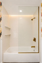 Minimalist bathtub-shower in a modern bathroom with white subway tile, clear glass panel, brass fixtures, built-in shelves and floral wallpaper accents.