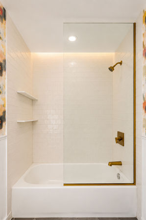 Minimalist bathtub-shower in a modern bathroom with white subway tile, clear glass panel, brass fixtures, built-in shelves and floral wallpaper accents.
