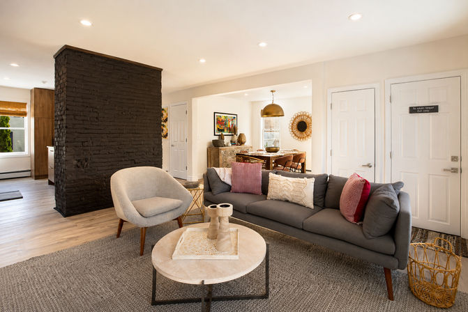 Cozy-chic open-plan living room and dining area with a gray mid-century sofa and textured pillows, curved knit armchair, round marble coffee table, dark painted brick divider, wood floors, woven accents and warm recessed lighting.