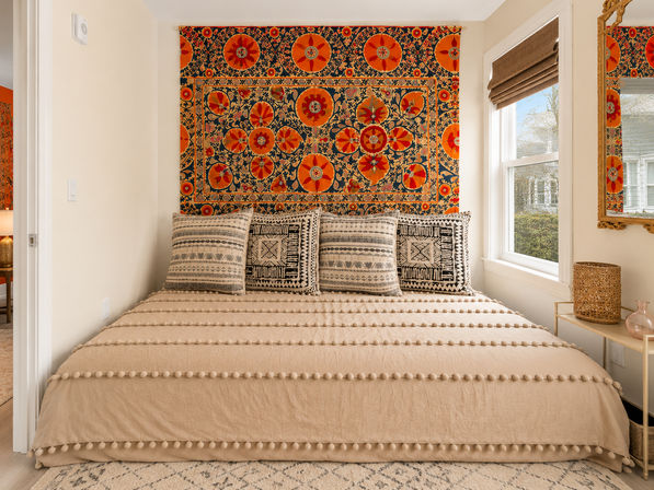 Cozy sunlit boho bedroom with a vibrant orange-and-navy floral tapestry above a bed dressed in a beige pom-pom duvet and patterned neutral throw pillows, woven side table and window with a roman shade.
