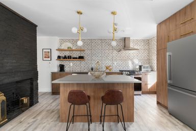 Modern open kitchen with wood cabinets and island topped with quartz, geometric tile backsplash, brass pendant lights and faucet, stainless steel fridge and range, leather bar stools, light wood flooring and black painted brick fireplace.