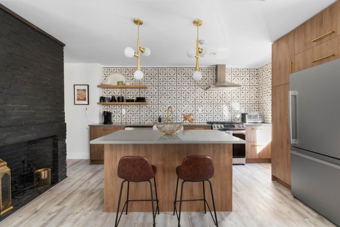 Modern open kitchen with wood cabinets and island topped with quartz, geometric tile backsplash, brass pendant lights and faucet, stainless steel fridge and range, leather bar stools, light wood flooring and black painted brick fireplace.
