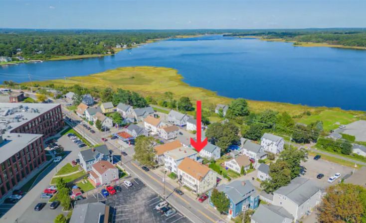 Aerial view of a coastal small‑town neighborhood with blue estuary and marshland, a red arrow highlighting a waterfront house