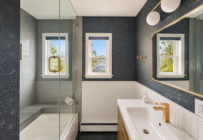 Bright modern bathroom with tub and glass shower, navy patterned wallpaper, white beadboard wainscoting, gold faucet and mirror trim, twin windows with leafy suburban view