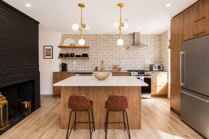 Bright modern kitchen with wood island and white quartz top, geometric tile backsplash, brass globe pendant lights, leather bar stools, stainless appliances and black painted brick fireplace