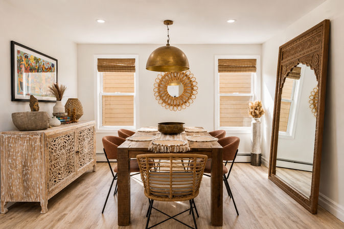 Cozy sunlit boho dining room with a rustic wooden table, rattan and upholstered chairs, carved whitewashed sideboard, brass pendant light, round rattan wall mirror and oversized ornate floor mirror in warm neutral tones.