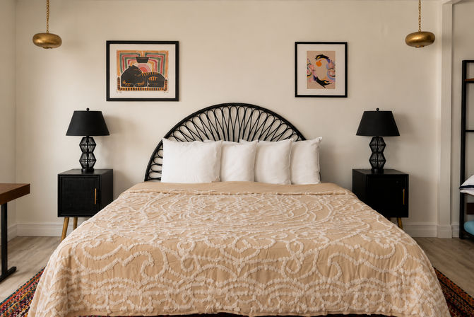 Contemporary boho bedroom with a king bed, black woven semicircle headboard, beige textured quilt and four white pillows, black nightstands with geometric lamps, framed art and brass pendant lights.