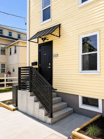 Cheerful yellow townhouse entrance with a black door, sloped black metal awning, concrete steps and modern horizontal black railing against yellow vinyl siding.