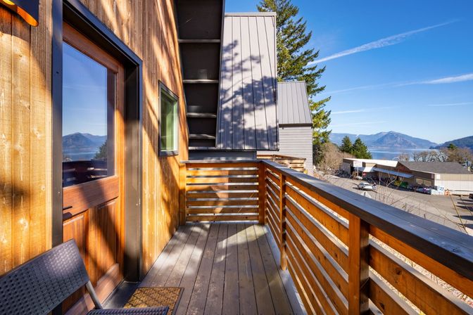 Sunny wooden balcony with slatted railing, offering an inviting view of a calm bay, forested mountains and a small coastal town under a clear blue sky.