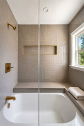 Sunlit modern bathroom with white bathtub and glass shower, beige herringbone tile accent wall, brass-gold showerhead and faucet, recessed brass-framed niche and a window showing green trees