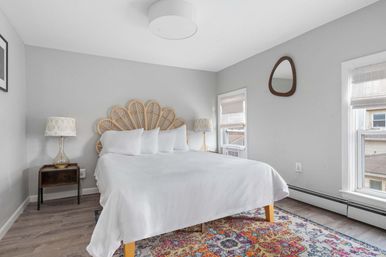 Bright, airy bedroom with a rattan fan-shaped headboard, crisp white bedding, glass bedside lamps, gray walls, two windows with woven shades, and a colorful patterned area rug.