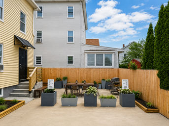 Sunny urban townhouse backyard patio with wooden picket fence, modern gray planters, outdoor dining table and grill between yellow and white siding homes.