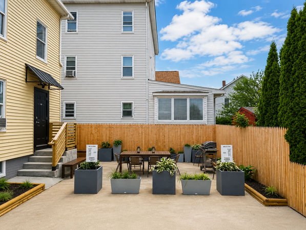 Sunny urban townhouse backyard patio with wooden picket fence, modern gray planters, outdoor dining table and grill between yellow and white siding homes.