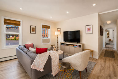 Cozy modern urban living room in an apartment — gray sofa with red accent pillows, cream armchair, TV on media console, floor lamp, bamboo blinds and light wood floors.