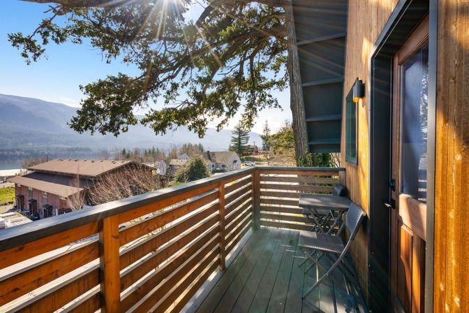 Sun-dappled wooden balcony with a small bistro set and railing, offering a lakeview across evergreen trees and distant mountains.