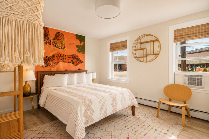 Cozy boho bedroom with white patterned bed, bold orange tiger mural above the headboard, macramé hanging, bamboo blinds, round gold wall shelf, wooden chair and sunlit windows.