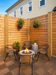 Cozy urban patio corner with a round fire pit, four woven rattan chairs, hanging planters and a tall horizontal wooden privacy fence on wood-tile decking