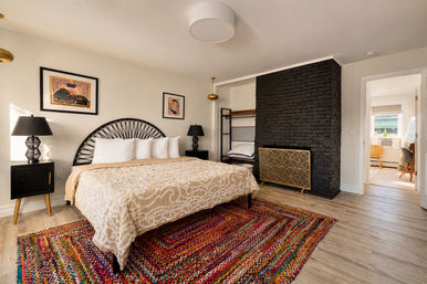 Bright boho apartment bedroom with beige patterned bedspread, black rattan headboard, black brick fireplace, colorful woven rug, bedside lamps, and open doorway to a sunlit workspace.