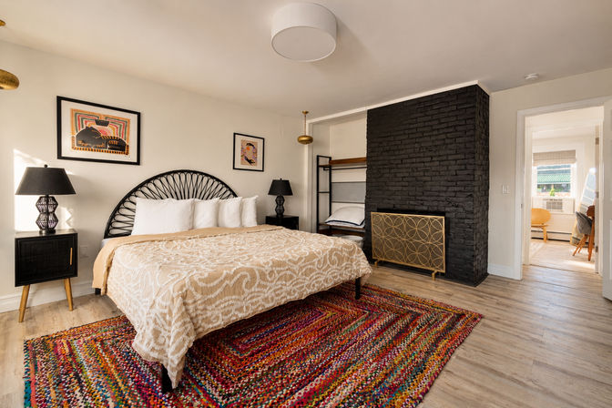 Bright boho apartment bedroom with beige patterned bedspread, black rattan headboard, black brick fireplace, colorful woven rug, bedside lamps, and open doorway to a sunlit workspace.